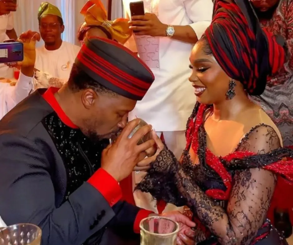 Sharon ooja and her husband in matching wedding attire. Sharon's outfit is made of red and black vertical patterned aso oke with lace detailing and he husband is in black cotton trad with a red and black cap to match her outfit